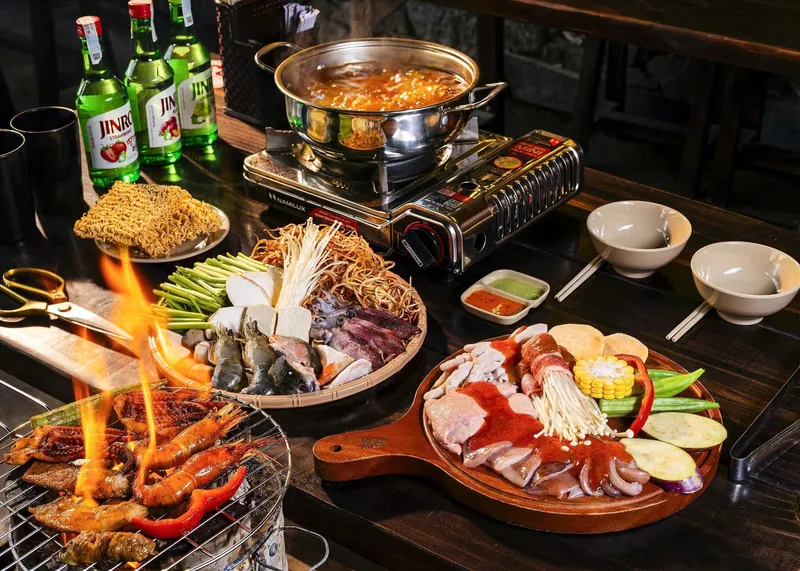 Bàn tiệc có nồi lẩu, lò nướng than đang cháy lửa với tôm mực, mâm đồ nhúng và khay thịt ba chỉ